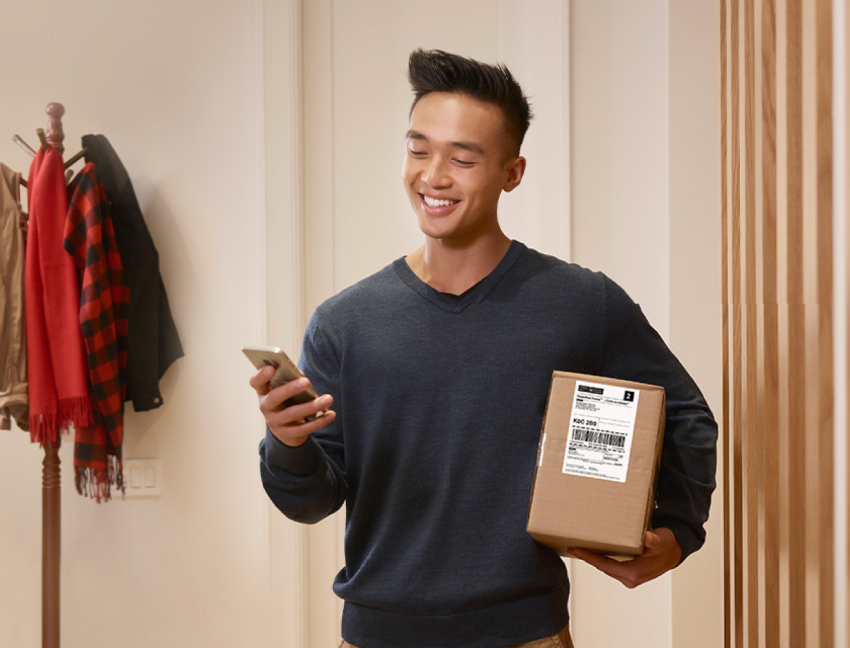 A smiling man looks at his smartphone as he holds a package.