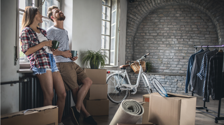 A man and a woman lean back against a windowsill in an apartment filled with moving boxes. 