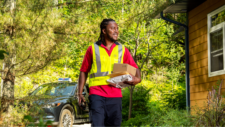 A mail carrier in a safety vest holds multiple parcels and walks through a treed residential area.