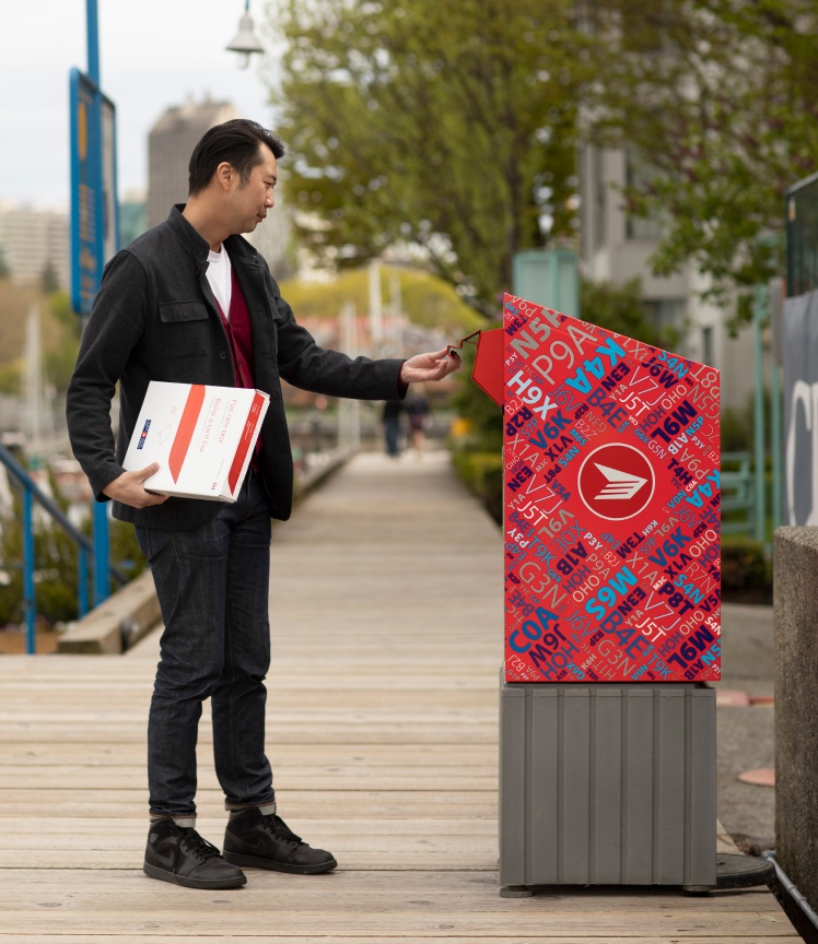 A man deposits a Canada Post flat rate box into a red Canada Post mailbox.