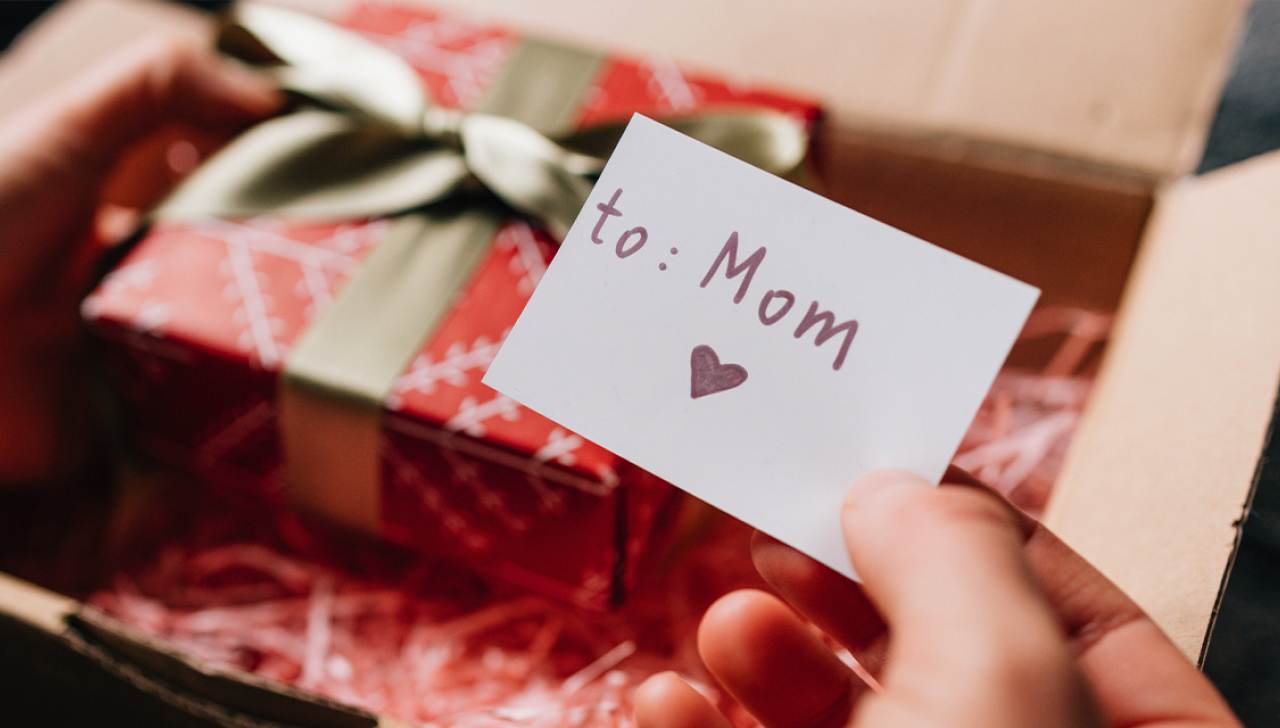 Hands open a shipping box which contains a wrapped gift box and a card that reads 'to Mom.’