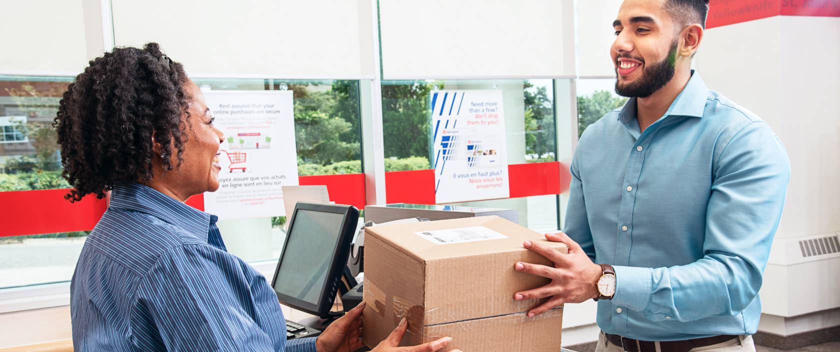 A smiling Canada Post retail employee hands a package to a man from behind a post office counter.