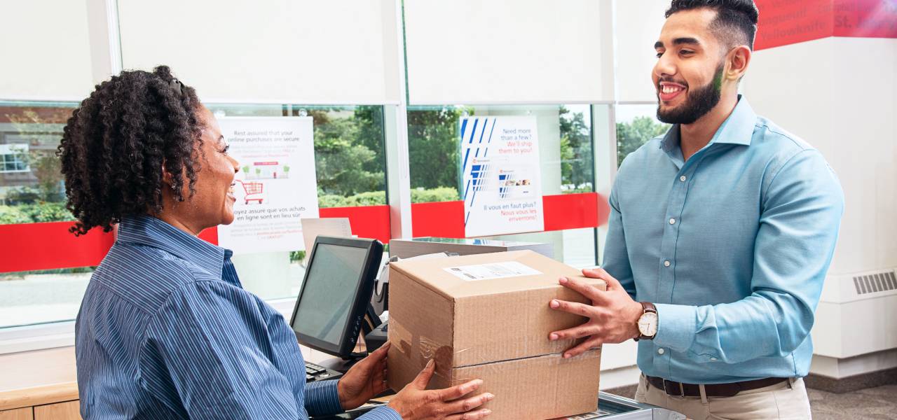 A smiling Canada Post retail employee hands a package to a man from behind a post office counter.