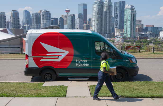 Une camionnette hybride de Postes Canada stationnée sur la rue avec en arrière plan, la silhouette du centre-ville de Calgary. Sur le trottoir, une employée de Postes Canada transporte un colis dans ses mains.