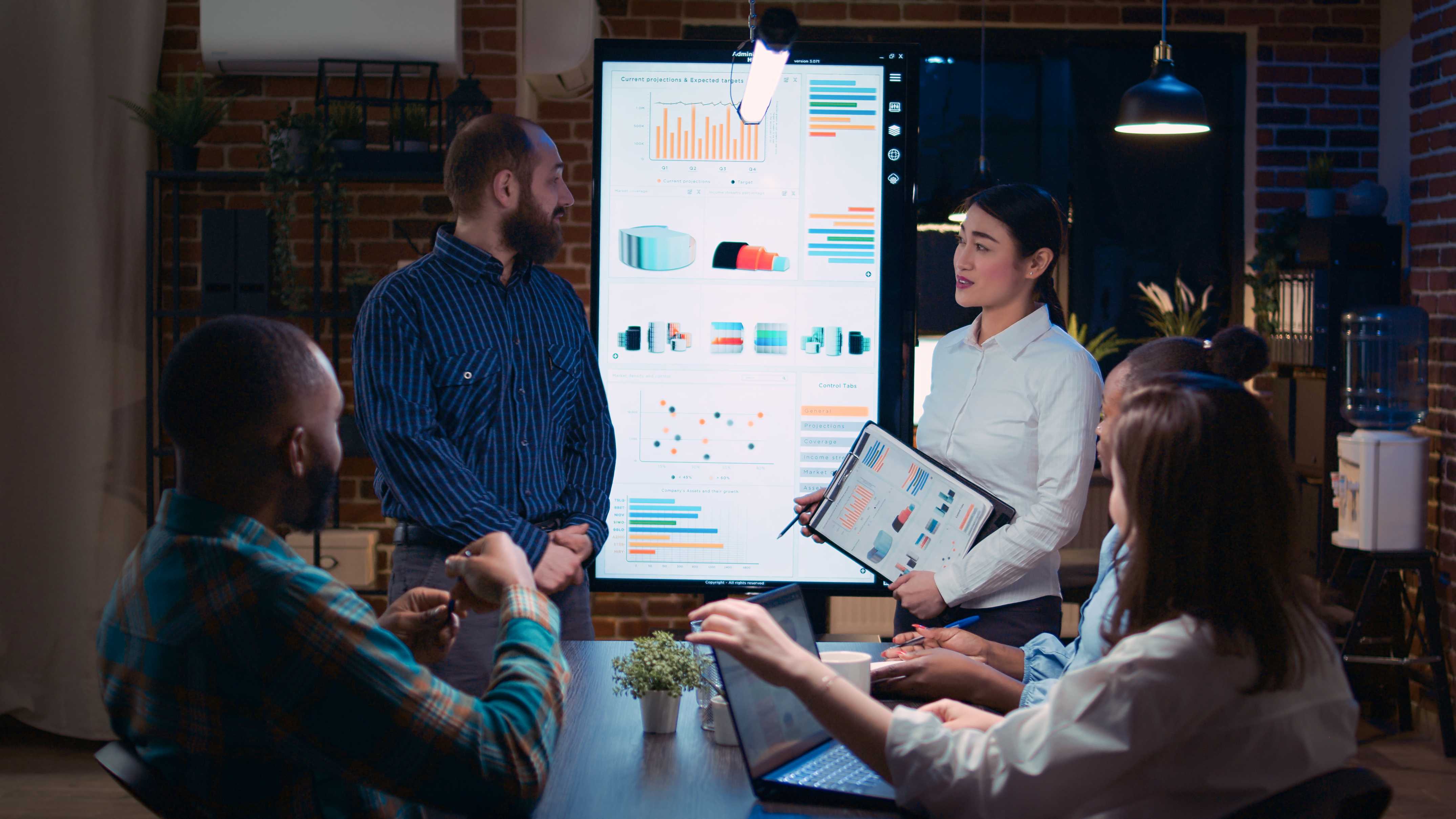 Deux personnes présentent des tableaux de données projetés à un écran durant une réunion.
