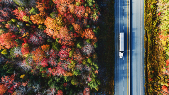 A white truck travels down a rural highway surrounded by dense and colourful trees in the fall.